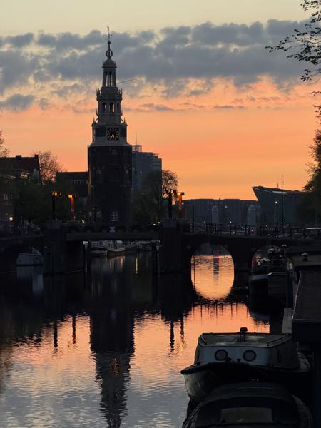 This captivating photo captures a stunning sunset over the Oudeschans canal in Uilenburg, Amsterdam, with the iconic Montelbaanstoren tower silhouetted against the vibrant orange sky